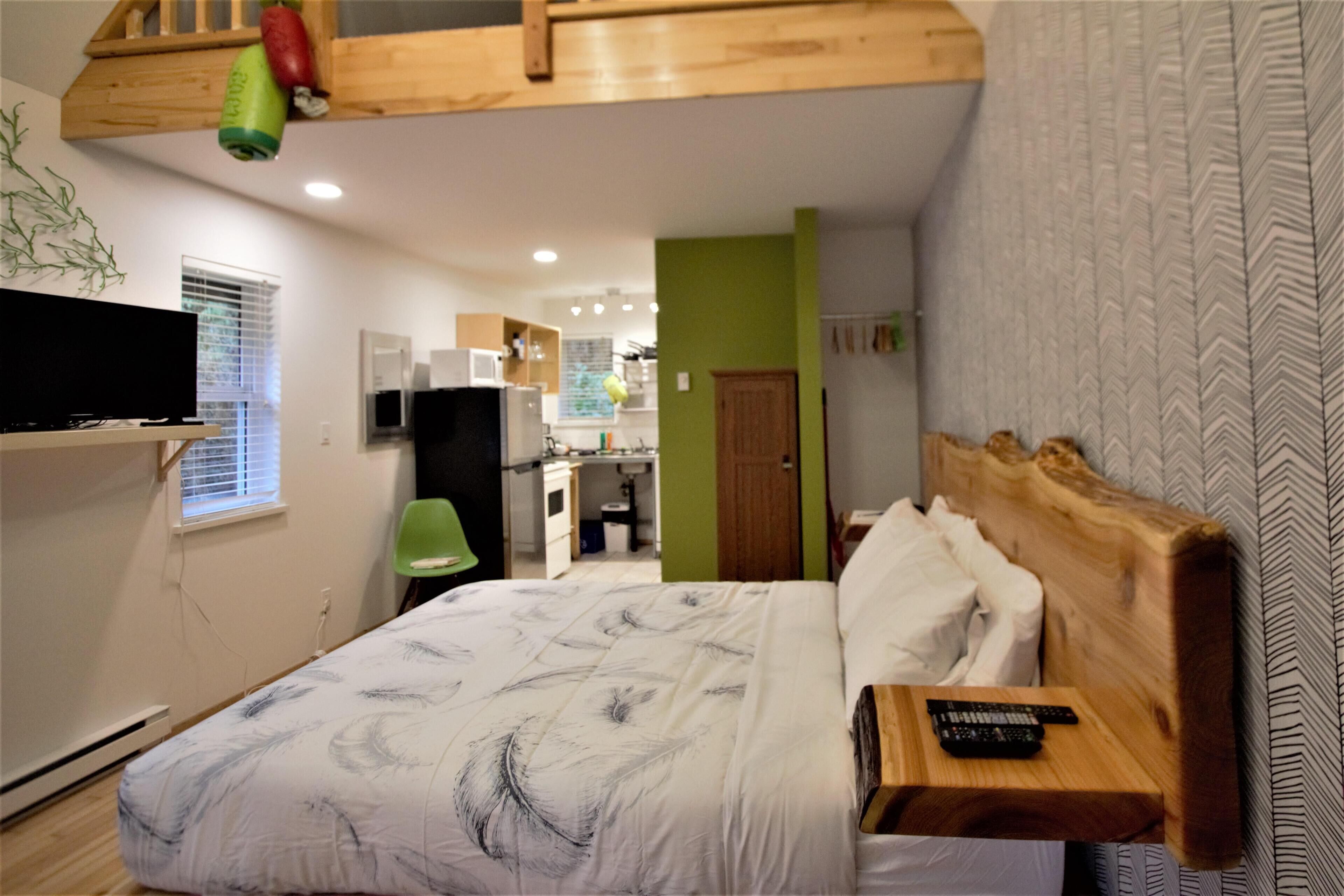 A bedroom with a wooden headboard, a bed with a feather-patterned duvet, a mounted TV, and a kitchenette in the background.