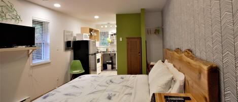 A bedroom with a wooden headboard, a bed with a feather-patterned duvet, a mounted TV, and a kitchenette in the background.