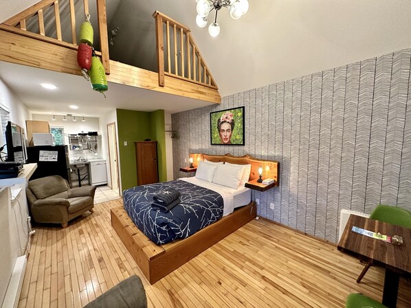 Un dormitorio con cama estilo loft, una pared verde, papel tapiz estampado, cabecera de madera y una mesita de noche pequeña con lámpara.