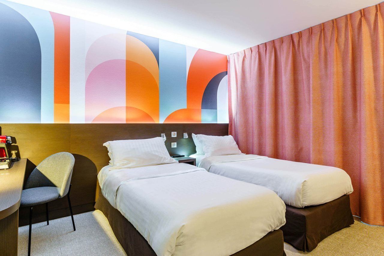 Une chambre d’hôtel avec deux lits, un bureau, une chaise et une fresque murale abstraite et colorée.