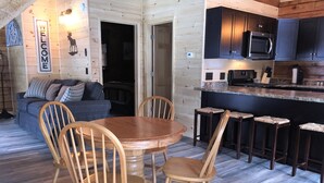 A cozy cabin interior with a wooden dining table and chairs, a kitchen area with dark cabinets and a microwave, and a living room with a sofa and decorative wall art.