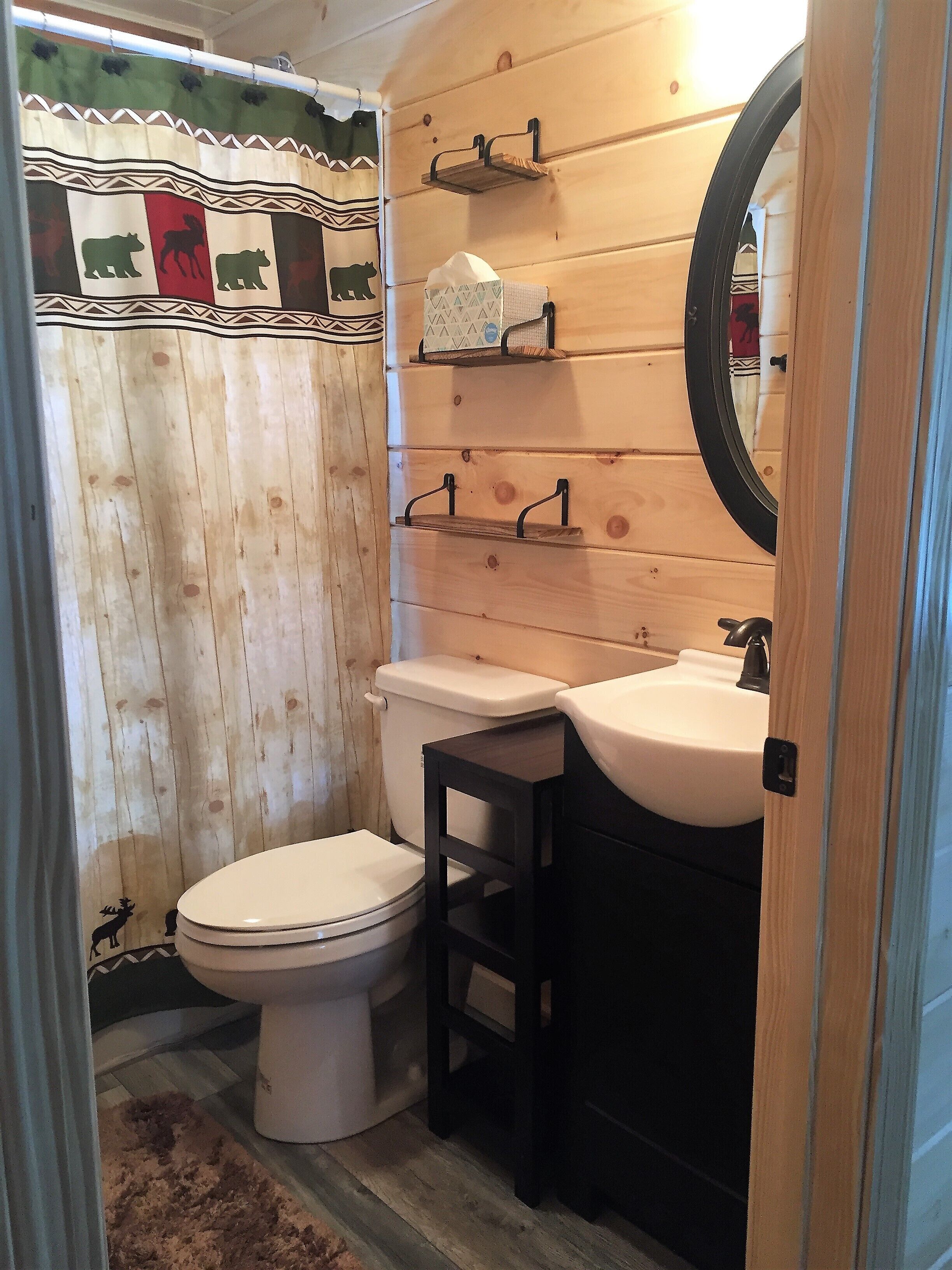 A bathroom with a wooden wall, a toilet, a sink, and a mirror. There are shelves above the sink and a towel hanging on the wall.