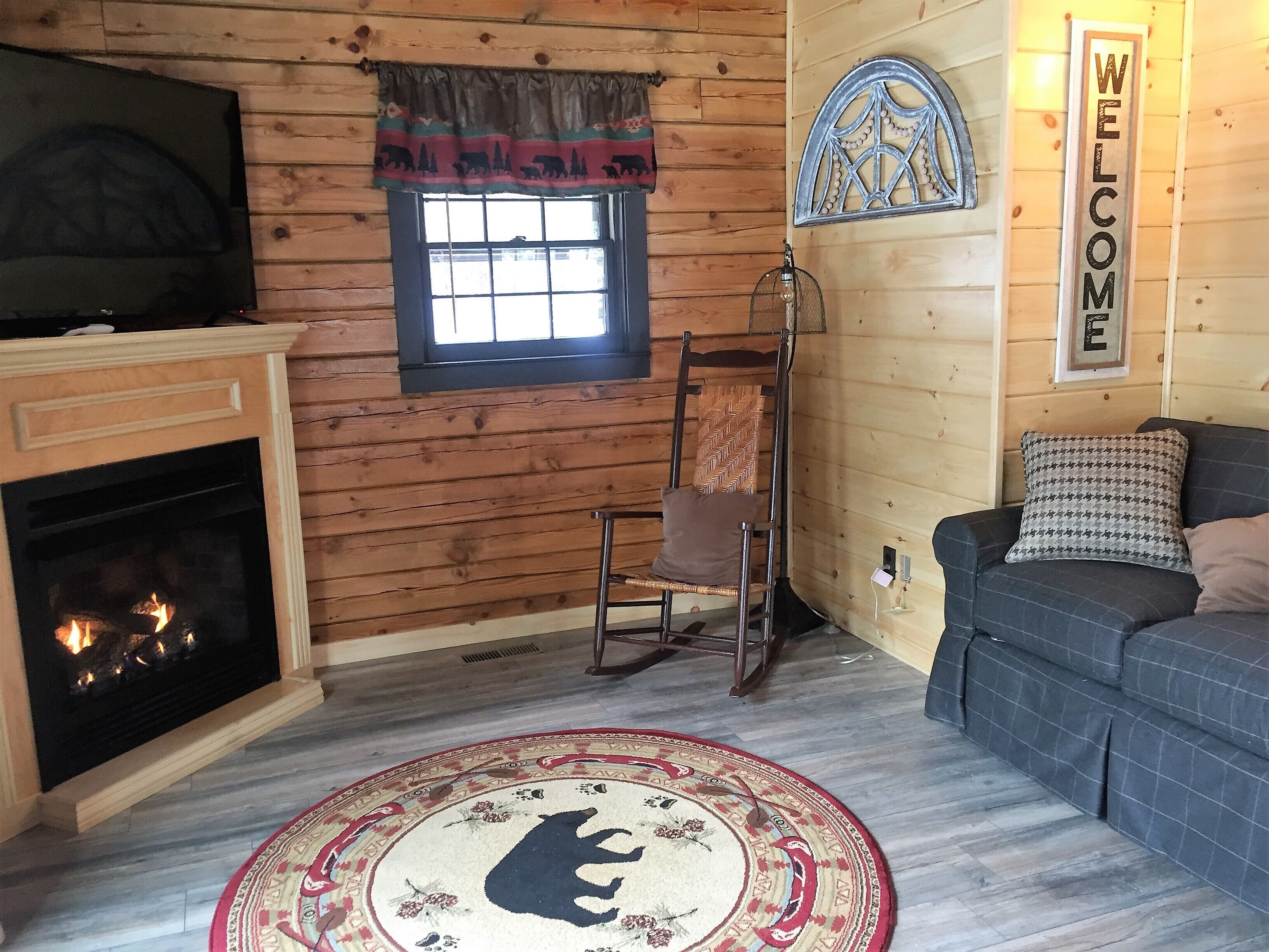 Ein gemütliches Blockhausschlafzimmer mit Kamin, Fernseher, einem 'WELCOME'-Schild, einem gemusterten Teppich und einer Couch.