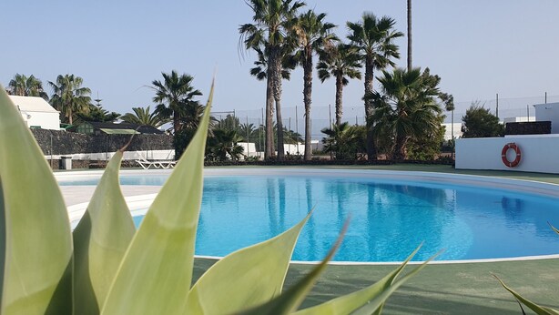 A swimming pool with a clear blue water, surrounded by palm trees and a white building. - Villa Las Palmeras in Playa Blanca (Playa Blanca)