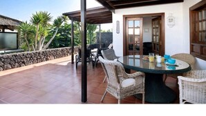 A covered patio with a round table and wicker chairs, a stone wall, and a view of palm trees. - Villa Las Palmeras in Playa Blanca (Playa Blanca)