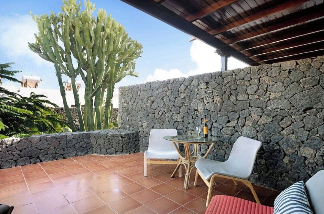 An outdoor patio with stone walls, two white chairs, a glass table, and a cactus in the background.