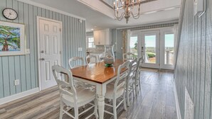 A dining room with a wooden table and white chairs, a chandelier, and a view of the outdoors. - Glad Tidings - Six Bedroom, Four Bath Inlet-Front Beach Home (Garden City Beach)