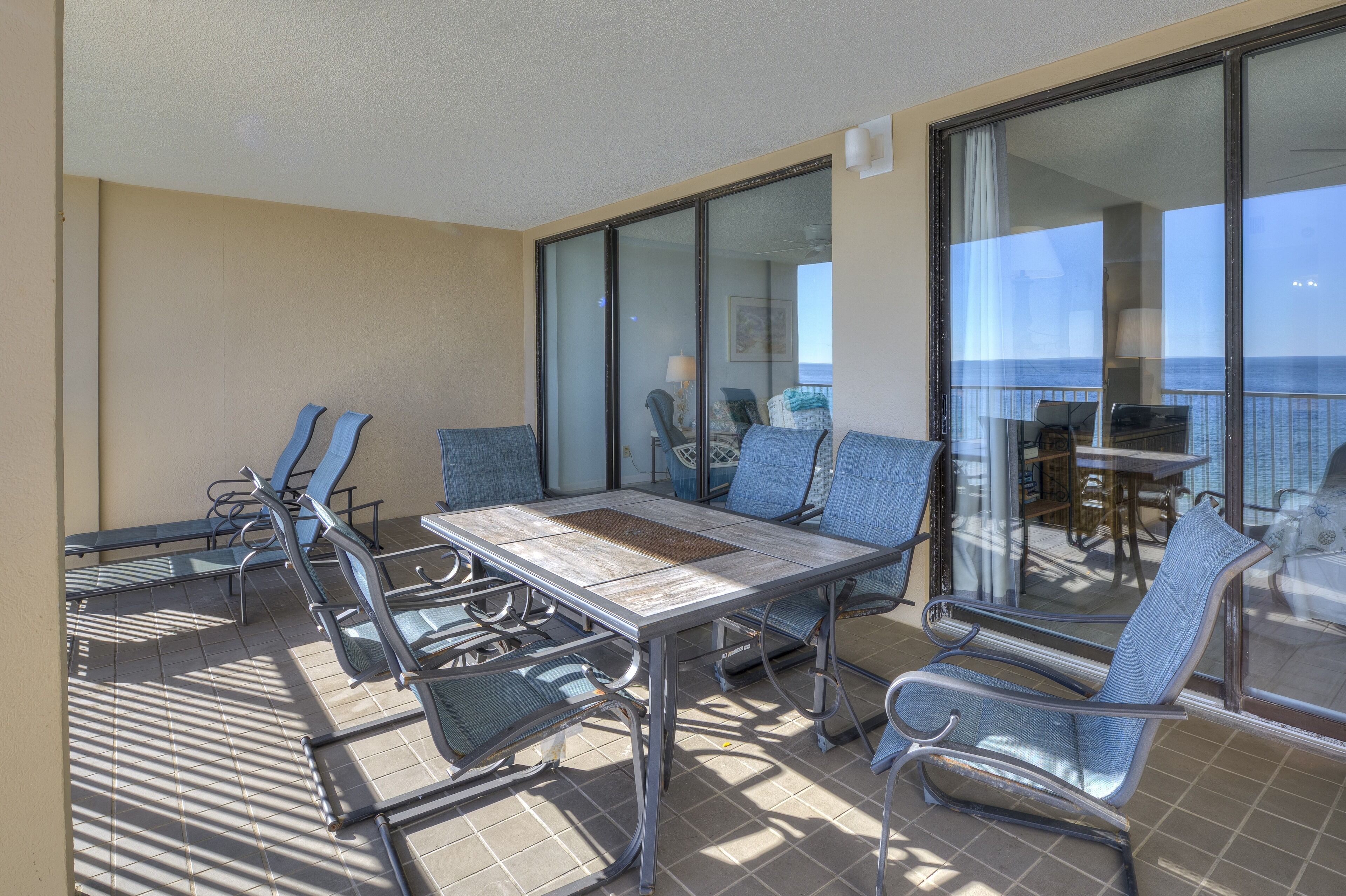 A sunlit balcony with a table and chairs, large windows, and a view of the ocean.