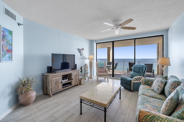 A spacious living room with a television, a coffee table, a sofa, and a balcony with a view of the ocean.