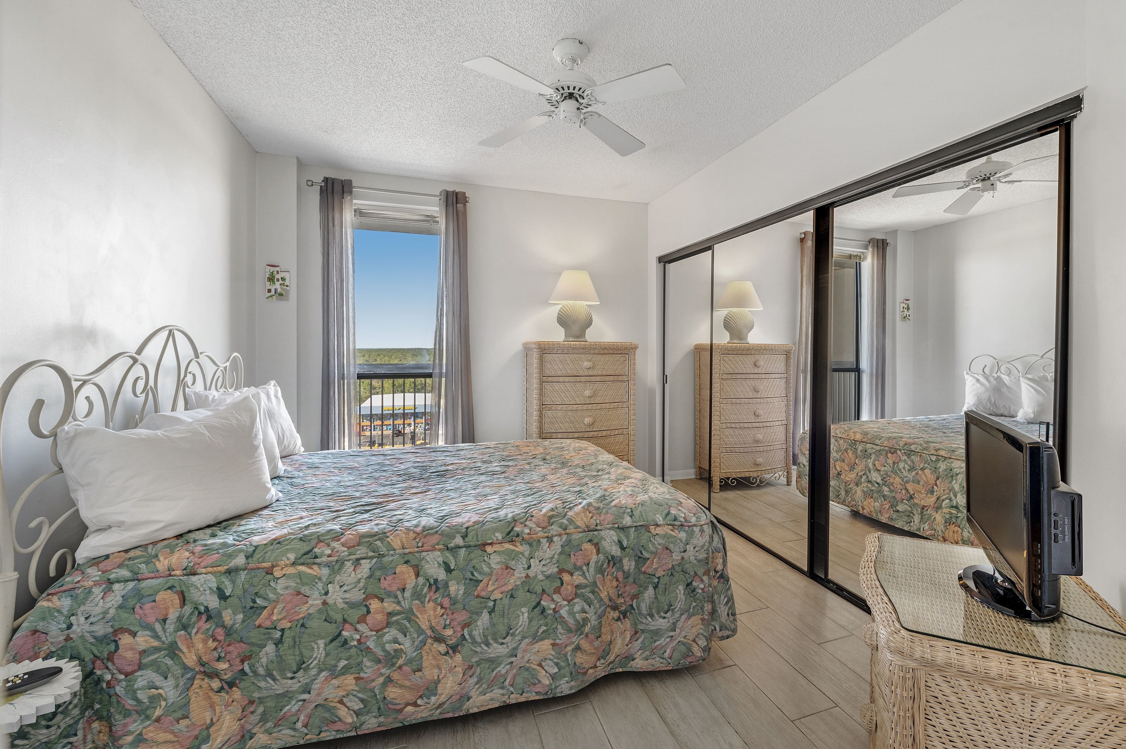 A bedroom with a bed, a dresser, a television on a side table, and a balcony with a view.