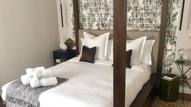 Un lit bien fait, avec du linge de lit blanc et un couvre-lit à motifs gris, une table de chevet avec un livre et une lampe, et un mur orné d’un motif abstrait en noir et blanc.