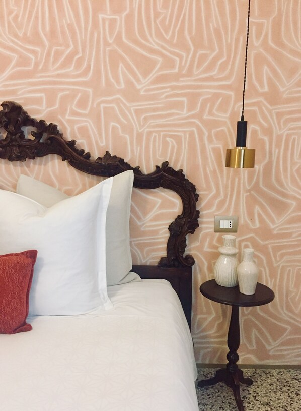 A neatly made bed with white linens and a red pillow, a wooden headboard, a small round table with two white vases, and a wall with a geometric pattern. - Addler House Apartment (Venezia)