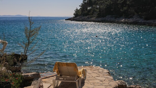 A chair by a rocky shoreline with a clear view of the sea and a forested hill in the background. - Secluded vacation home by the sea (Milna)