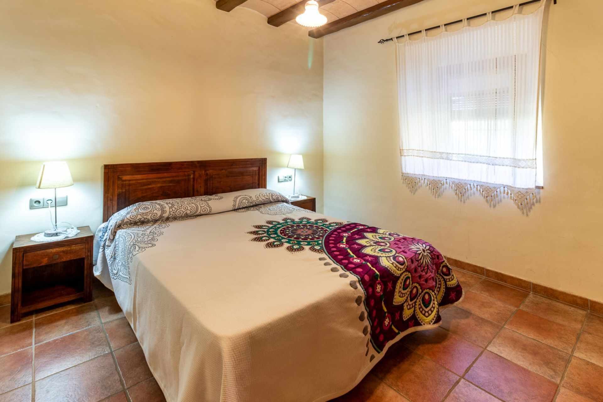 A bedroom with a large bed, two bedside tables with lamps, a window with sheer curtains, and a wooden ceiling beam.