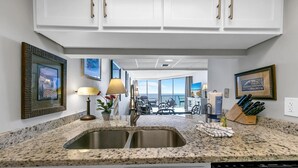 A kitchen with granite countertops, stainless steel sinks, and white cabinetry. There are framed pictures on the walls, a potted plant, and a knife block on the counter.