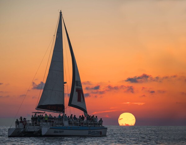 Un velero con pasajeros navegando al atardecer, con el sol visible al fondo.