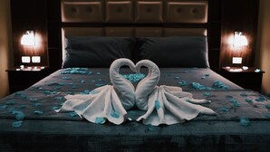 A bed with a heart-shaped towel arrangement and rose petals, flanked by two bedside lamps.
