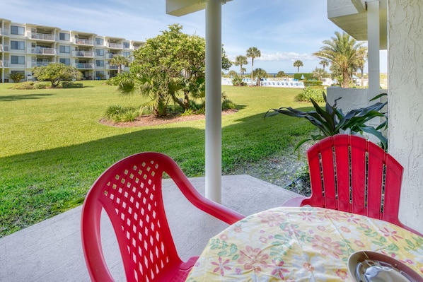 A patio with two red chairs and a table, overlooking a well-maintained lawn and a building with multiple balconies.