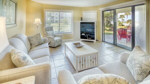 A living room with a sofa, armchairs, a coffee table, a TV, and a sliding glass door leading to an outdoor area with chairs. - CRC 1111 - Beautiful Ground Floor Pool View Condo (St Augustine)