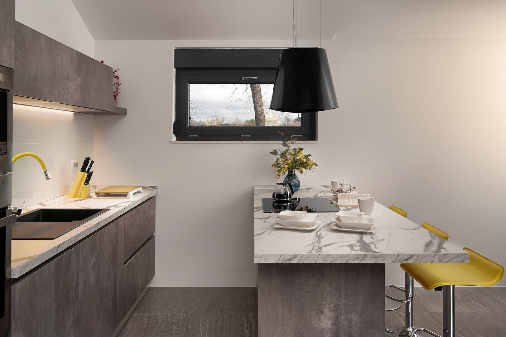 Une cuisine moderne avec un plan de travail en marbre, un luminaire suspendu noir et une vue sur l’extérieur par une fenêtre.