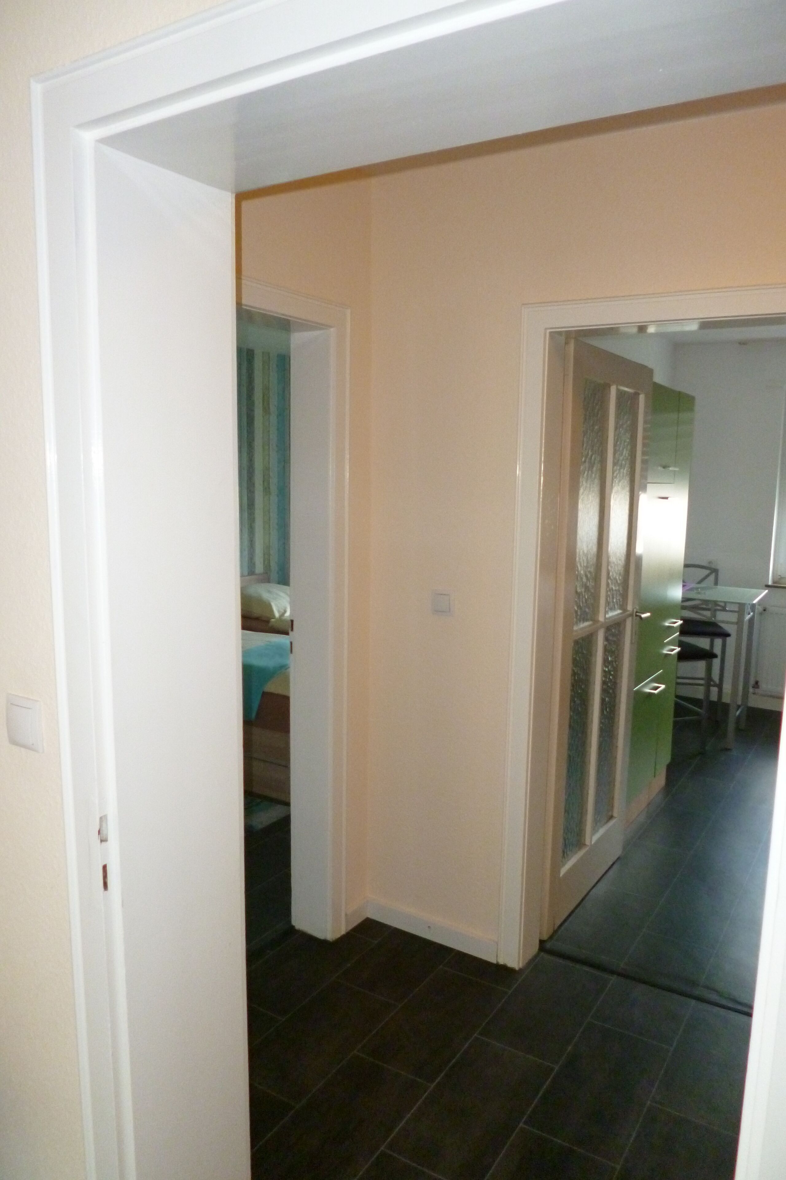 A hallway with a bed in the background, a kitchen area with a table and chairs, and a door leading to another room.