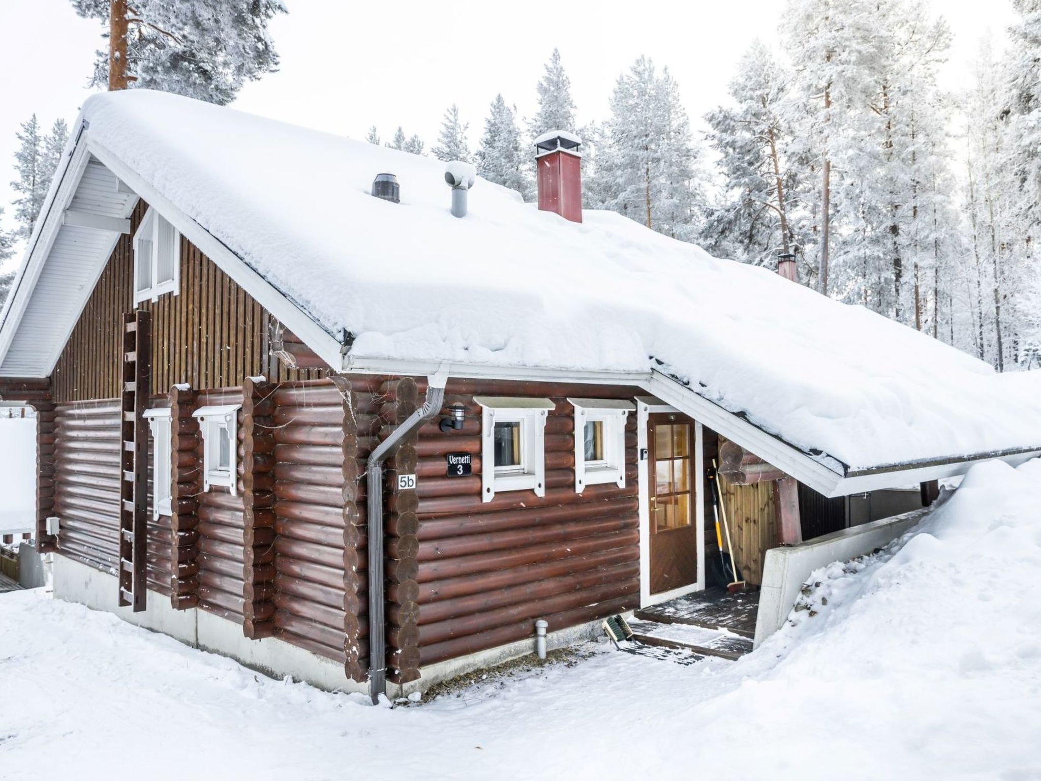 Une cabane en rondins avec un toit enneigé, une cheminée et un petit porche avec des marches menant à l’entrée.