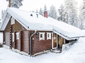 Une cabane en rondins avec un toit enneigé, une cheminée et un petit porche avec des marches menant à l’entrée.