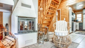 Un interior de cabaña acogedor con chimenea, paredes de madera, una zona de comedor con una silla de mimbre y una cocina visible a través de una puerta abierta.
