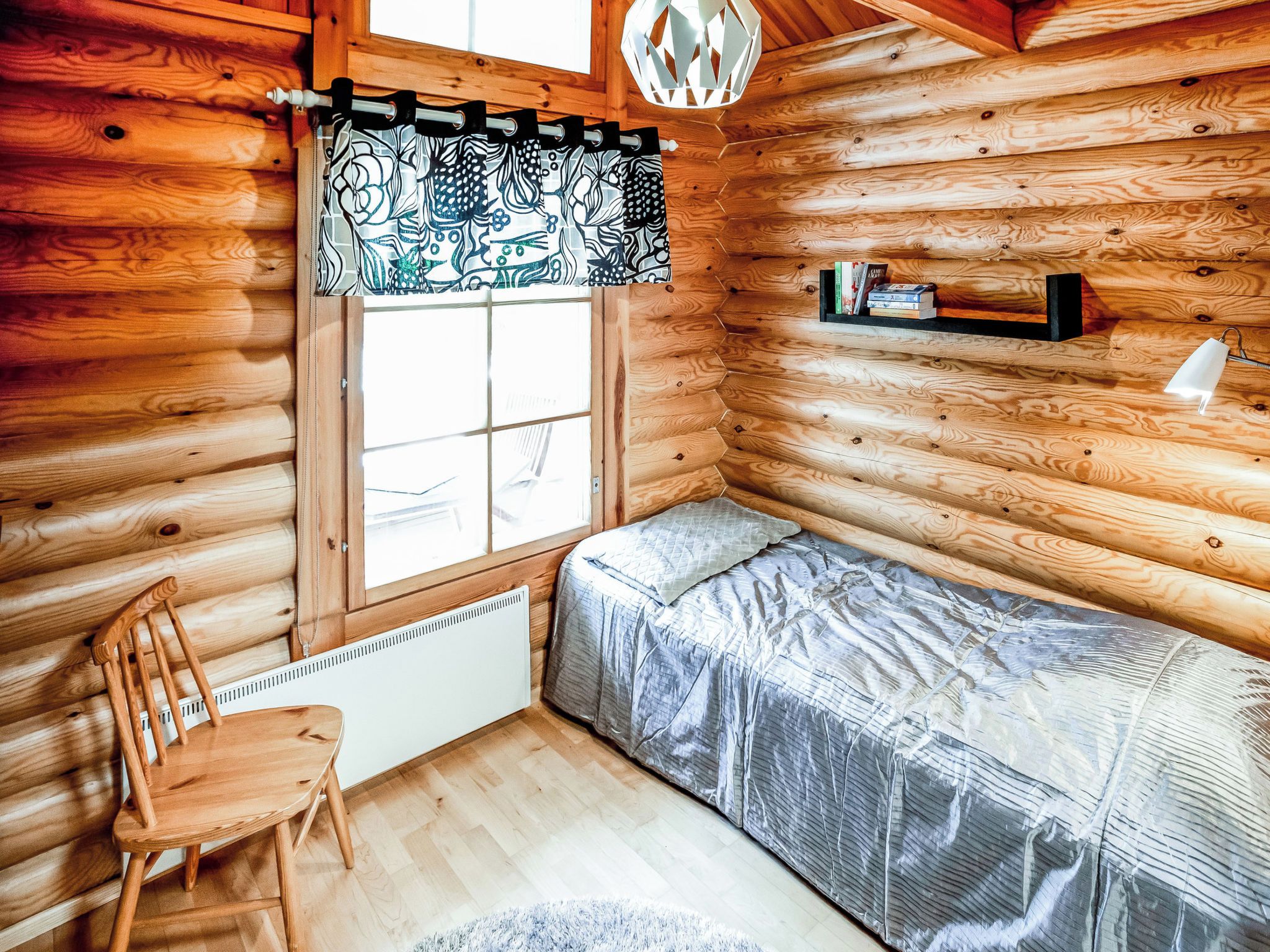 A cozy cabin room with a wooden interior, a window with a patterned curtain, a bed with a gray cover, a chair, a shelf with books, and a white radiator.