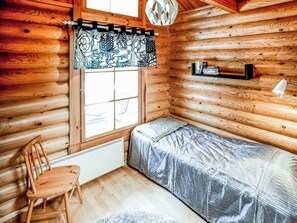 A cozy cabin room with a wooden interior, a window with a patterned curtain, a bed with a gray cover, a chair, a shelf with books, and a white radiator. - Kolin vernetti 3 by Interhome (Lieksa)