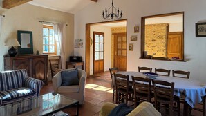 A spacious living room with a high ceiling, wooden beams, a chandelier, a dining area with a table and chairs, a sofa, a chair, a television, and a window with curtains. - In a green setting, pretty farm (producing walnuts and strawberries), swimming pool (Cendrieux)