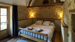 A bedroom with a stone wall, wooden beams, a bed with pillows, and a window with curtains. - In a green setting, pretty farm (producing walnuts and strawberries), swimming pool (Cendrieux)