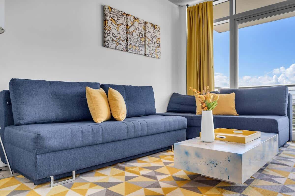 Ein modernes Wohnzimmer mit einem blauen Sofa, einem weißen Couchtisch und einem gelben Teppich mit Muster.