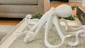 Una sala de estar con un sofá color beige, una escultura de pulpo blanco sobre una alfombra texturizada, y una cocina al fondo.
