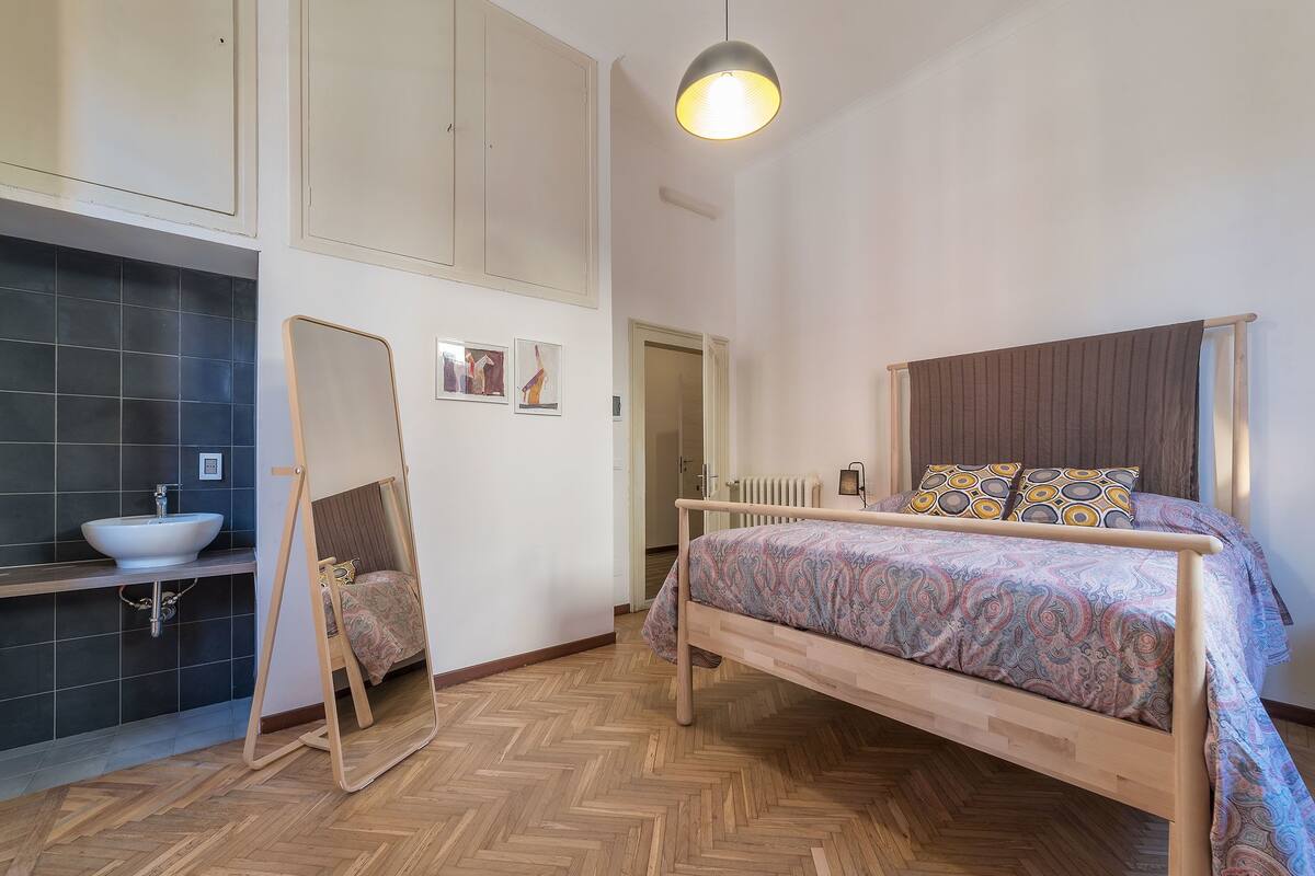 A bedroom with a bed, a bedside table, a mirror, a radiator, and a bathroom with a sink and tiles.