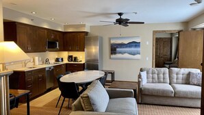 A modern kitchen with dark wood cabinets, a dining table, and a sofa in a living area with a ceiling fan. - Peaks Penthouse #643 (Mountain Village)