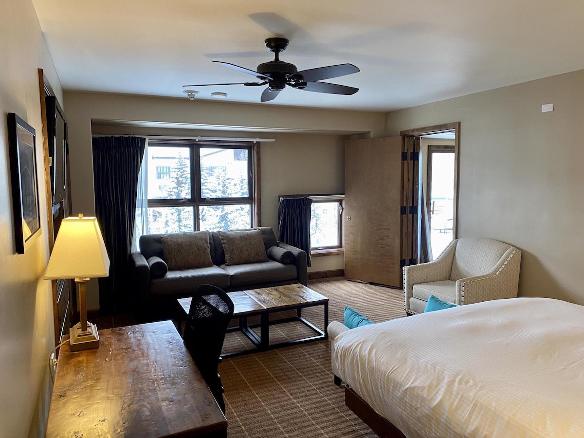 Quarto de hotel com cama, sofá, poltrona, mesa de centro e escrivaninha com abajur.