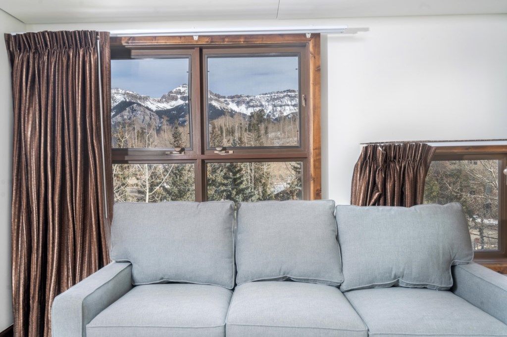 A room with a grey sofa facing a window with a mountain view.