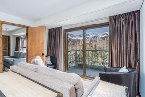Een hotelkamer met een bed, een bank, een stoel en een grote glazen schuifdeur naar een balkon met uitzicht op de bergen.