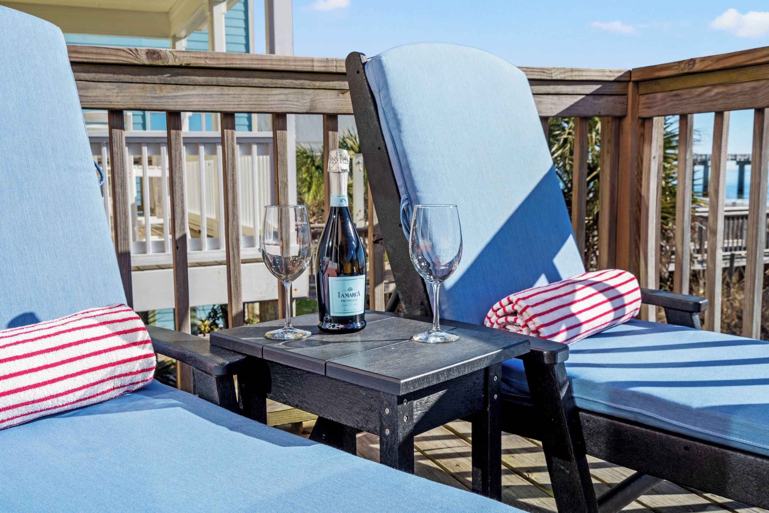 A wooden deck with two blue lounge chairs, each with a red and white striped pillow. A bottle of wine and two wine glasses are placed on a small table between the chairs.