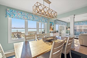 Un comedor con mesa y sillas de madera, un candelabro y ventanas amplias con vista al océano.