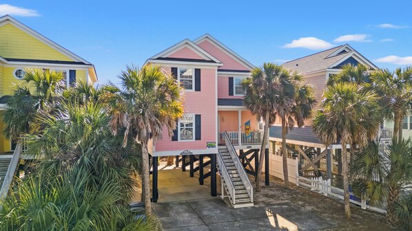 Una hilera de casas de playa, con una casa central de color rosa que tiene un balcón y una escalera que lleva a un porche cubierto.