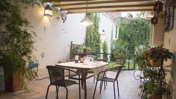 Un patio al aire libre con una mesa redonda y cuatro sillas, rodeado de macetas con plantas y un muro de ladrillo.