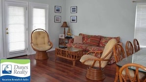 Ein Wohnzimmer mit einer Couch, zwei Sesseln, einem Couchtisch und einem Esstisch mit Stühlen. An der Wand befinden sich gerahmte Bilder, eine Lampe auf einem Beistelltisch und eine Tür mit Jalousien.