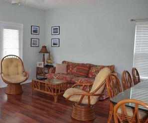 A living room with a sofa, two armchairs, a coffee table, and a dining table with chairs. There are framed pictures on the wall, a lamp on a side table, and a door with blinds.