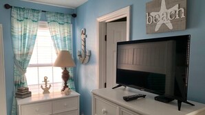 A room with a television on a white dresser, a lamp, and a window with curtains.