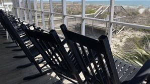 Uma área de estar coberta ao ar livre, com bancos de madeira e grandes janelas com vista para uma praia com passarelas de madeira.