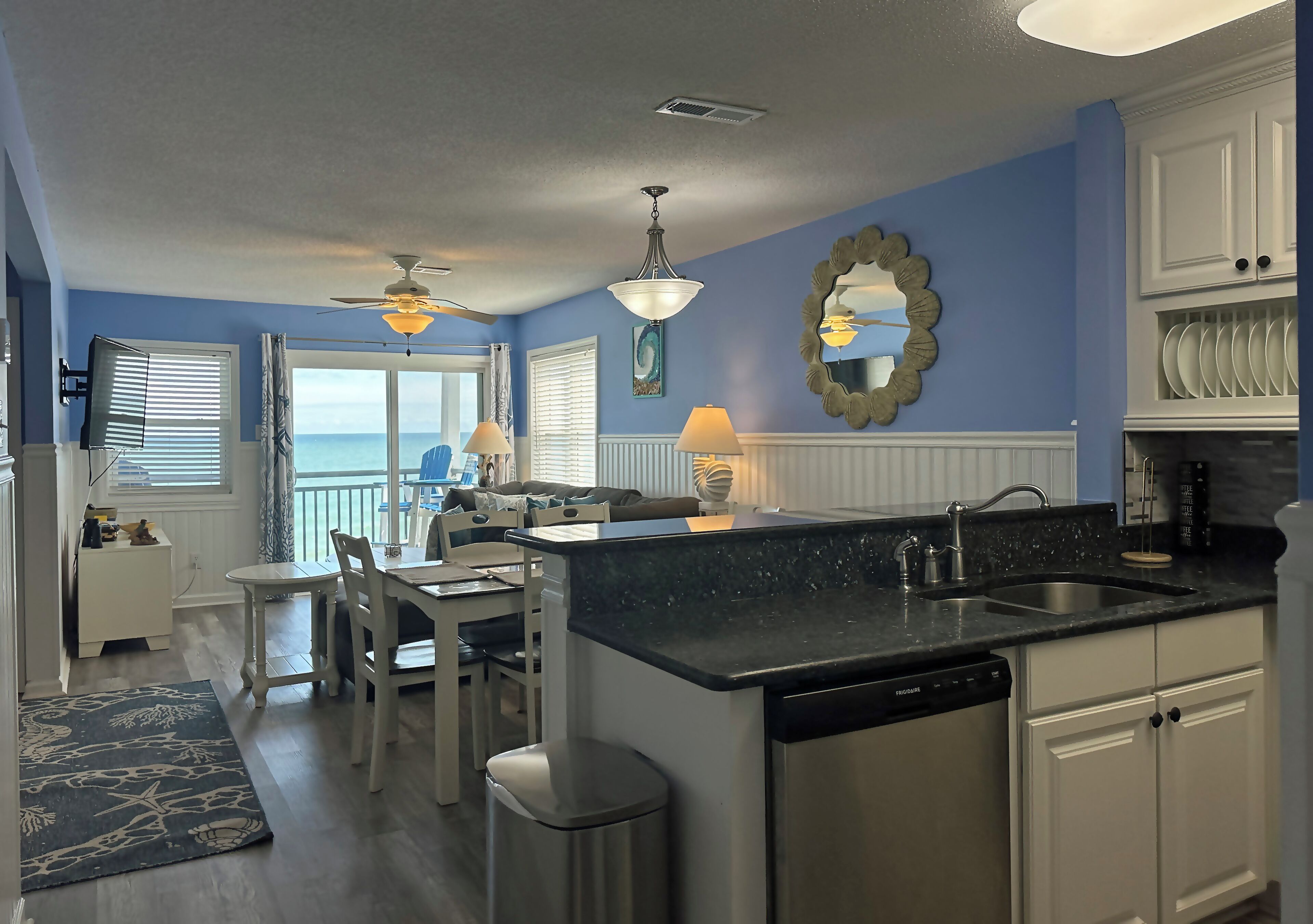 A kitchen with a black countertop, white cabinets, and a dishwasher. There is a dining area with a table and chairs, and a view of the ocean through the sliding glass door.