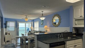 A kitchen with a black countertop, white cabinets, and a dishwasher. There is a dining area with a table and chairs, and a view of the ocean through the sliding glass door. - Dolphin Cove 3A - Oceanfront Condominium in Garden City Beach (Garden City)
