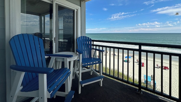 A balcony with two blue Adirondack chairs and a small table, overlooking a beach with umbrellas and people. - Dolphin Cove 3A - Oceanfront Condominium in Garden City Beach (Garden City)
